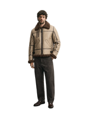 A man wearing a beige aviator-style jacket with dark brown faux-fur trim, paired with dark brown trousers and brown leather shoes, standing against a light gray studio background.