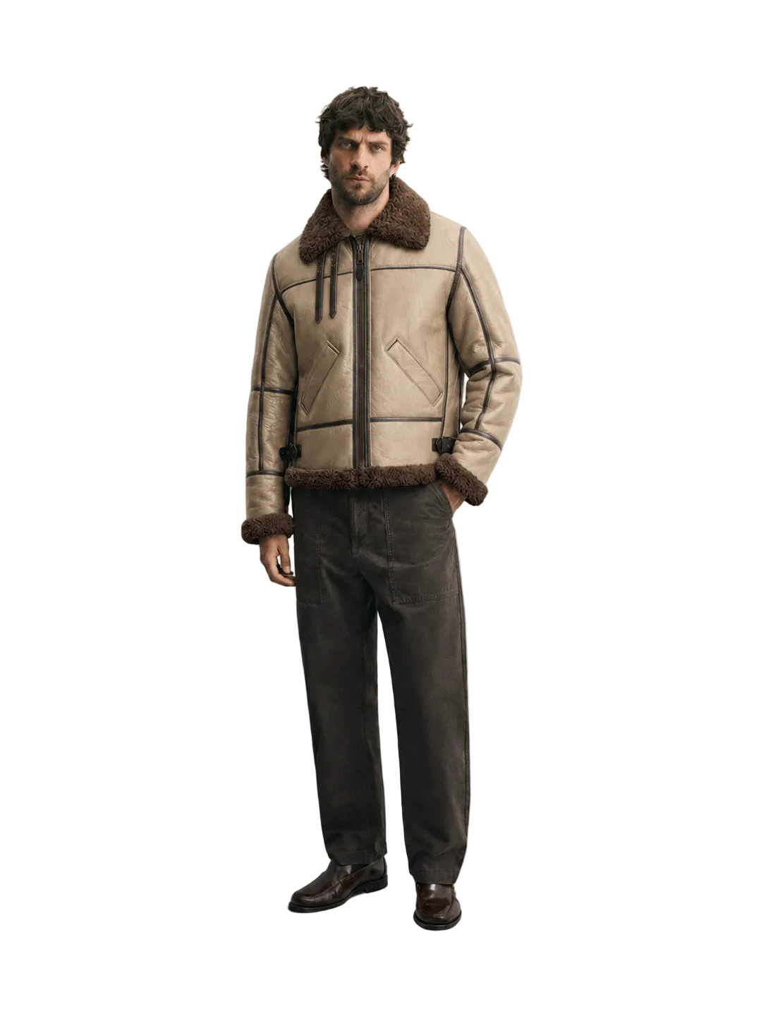 A man wearing a beige aviator-style jacket with dark brown faux-fur trim, paired with dark brown trousers and brown leather shoes, standing against a light gray studio background.