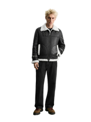 A model stands against a light gray studio background wearing a black leather aviator-style jacket with white shearling lining, black jeans, and brown shoes.