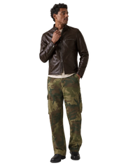 A man modeling a dark brown leather jacket and green camouflage cargo pants against a neutral gray studio background.