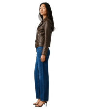 Woman in brown leather moto jacket, blue wide-leg jeans, and black heels, side view