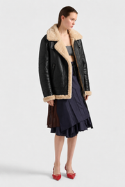 Woman wearing black shearling coat with cream fur lining, navy pleated skirt, and red heels