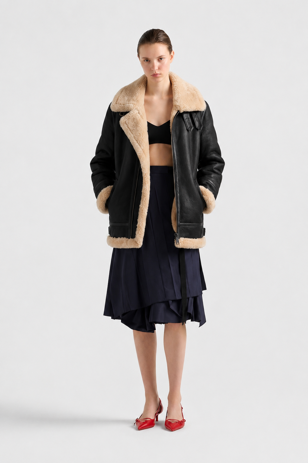 Woman in black shearling coat with tan fur lining, navy pleated skirt, red heels