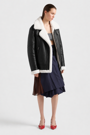Black shearling coat with white fur lining, worn over gray top and navy pleated skirt with red heels