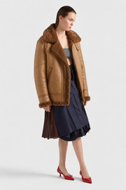 Women's brown shearling aviator jacket with plush collar, paired with navy pleated skirt and red heels