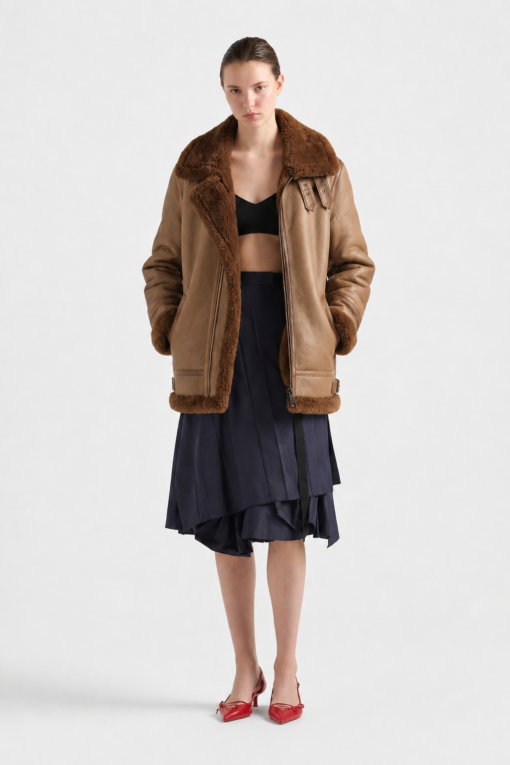 Brown shearling aviator jacket with fur collar, paired with navy pleated skirt and red heels
