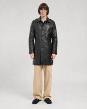 Men's black leather trench coat with button front and zipper pocket, styled with beige pants and loafers