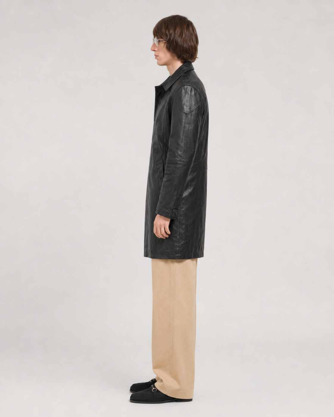 Man in black leather trench coat with beige pants and loafers, side profile