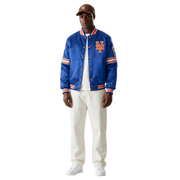 Men's royal blue Mets satin bomber jacket with orange trim and team logo, styled with white pants and cap