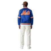Blue Mets varsity jacket with orange lettering, worn by man in white pants and cap, back view