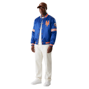 Men's royal blue New York Mets satin varsity jacket with orange trim and team logo, styled with white pants and cap