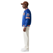 Man wearing blue New York Mets varsity jacket with orange trim, white pants, and brown cap