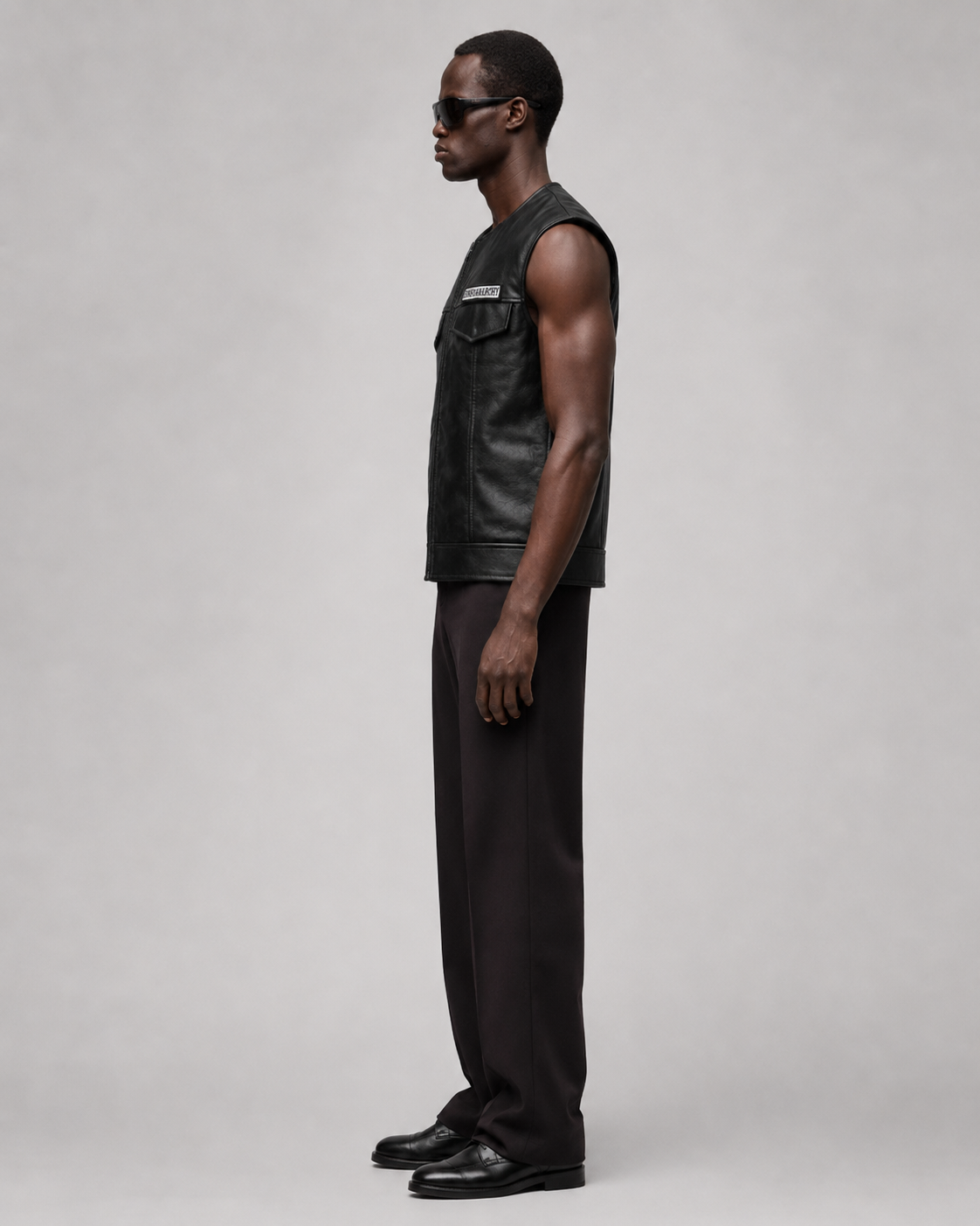 Men's black leather vest with patch, styled with dark trousers and sunglasses for a sleek, modern look.