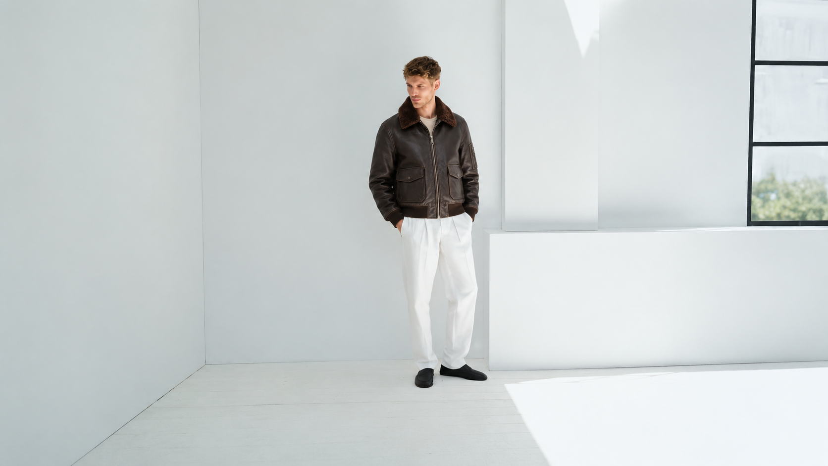 Man wearing brown leather bomber jacket with shearling collar, white pants, black shoes in minimalist room