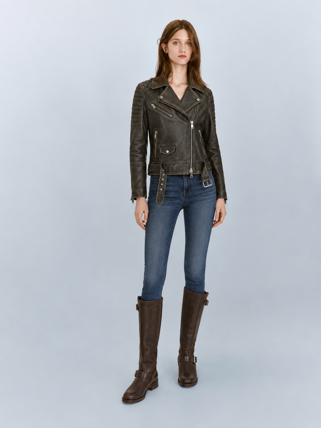 Woman in dark brown leather moto jacket, blue jeans, and knee-high boots on light background