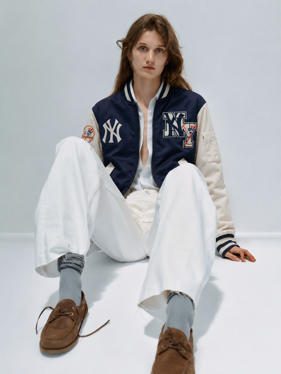 Woman wearing navy and cream varsity jacket with NY logo, white pants, gray socks, brown loafers