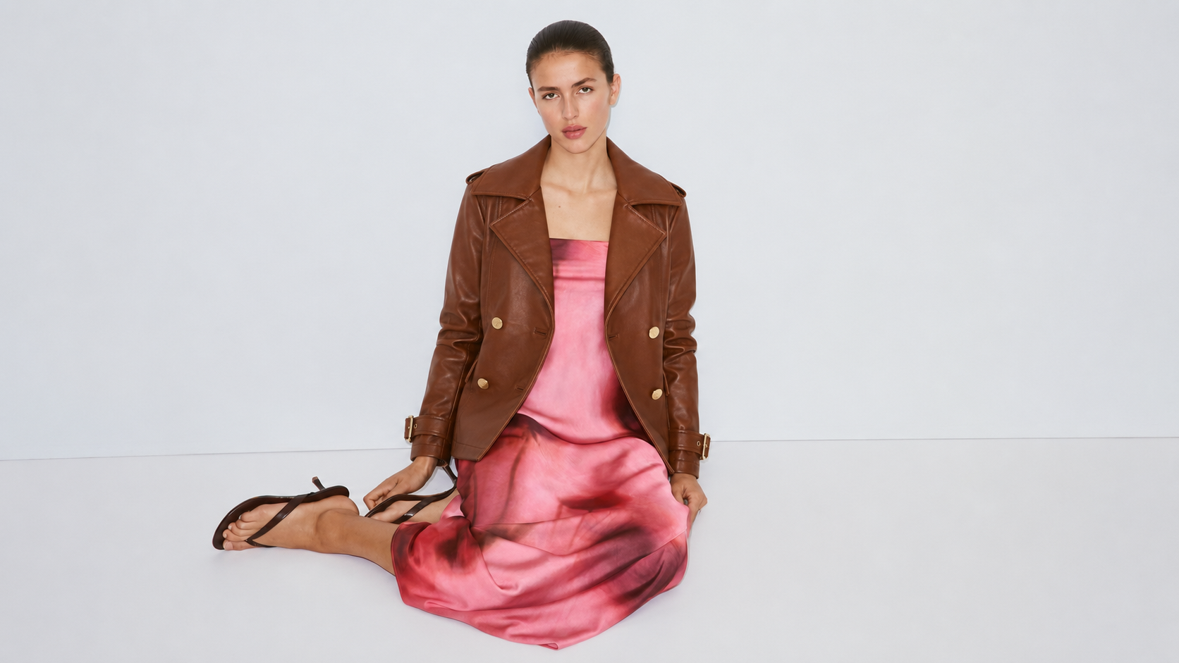 Woman in brown leather jacket and pink satin dress, sitting on white floor