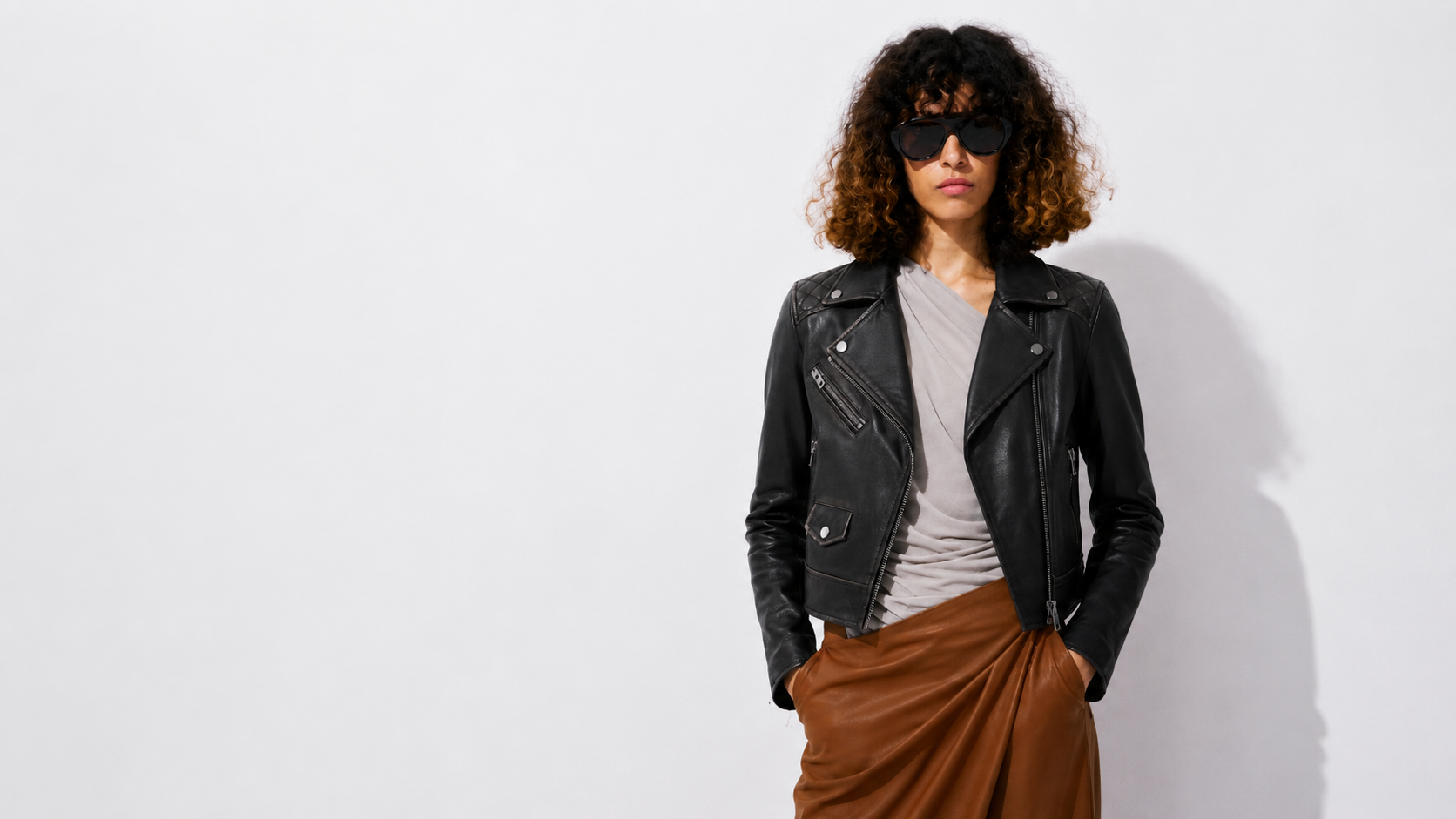Woman in black leather biker jacket, brown skirt, and sunglasses against white background