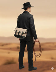 Man in black leather jacket and hat carrying beige satchel bag, holding rope in desert landscape
