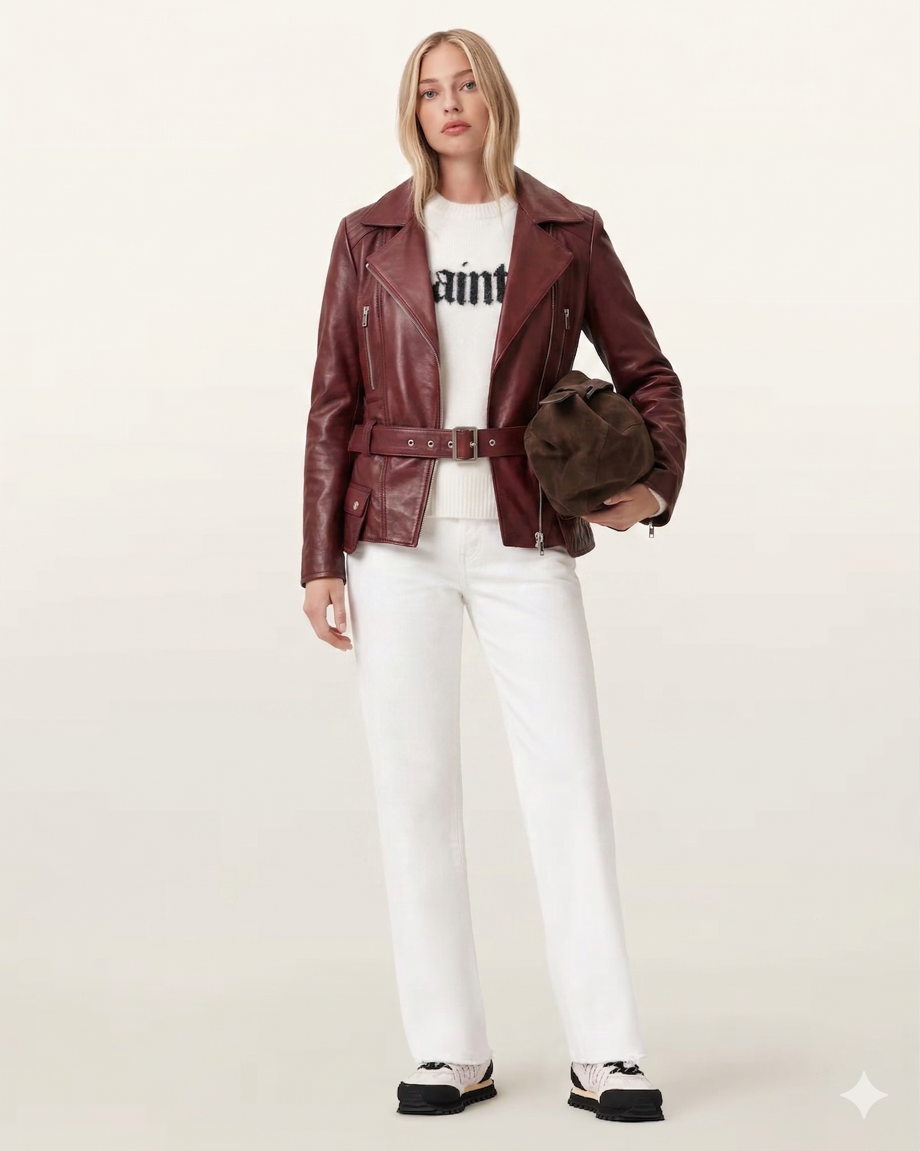 Woman in burgundy leather moto jacket, white jeans, and graphic sweater, holding brown bag