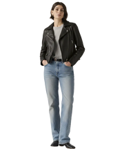 Woman wearing black leather jacket, gray tee, and blue jeans with hands in pockets
