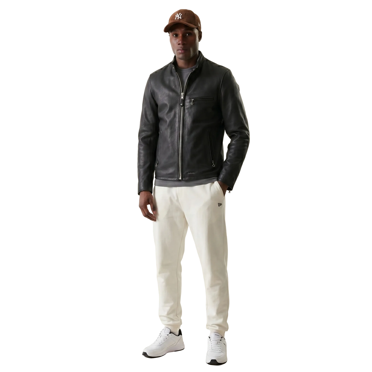 Man wearing a black leather jacket, white pants, and a brown cap on a beige background