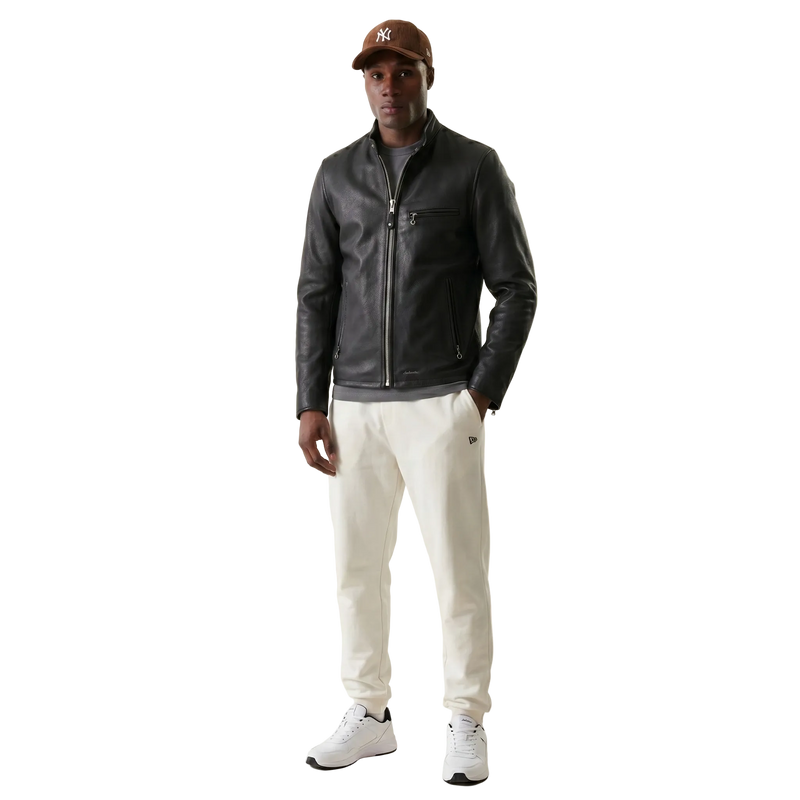 Man wearing a black leather jacket, white pants, and a brown cap on a beige background