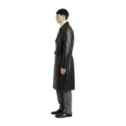 Man wearing black leather trench coat with belt, beanie, and gray trousers, side view