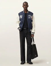 Man wearing navy and cream varsity jacket with NY logo, black jeans, and holding a tote bag
