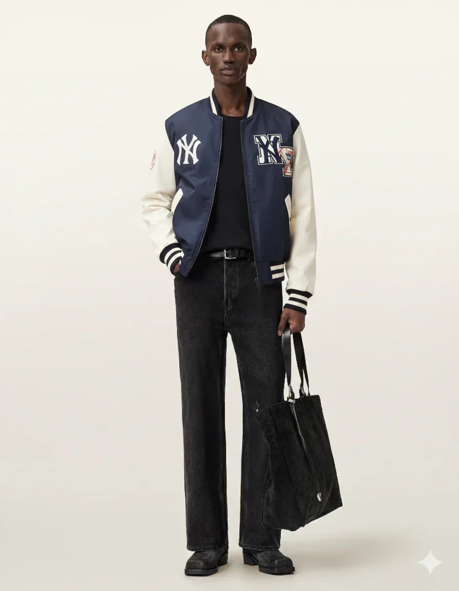 Man wearing navy and cream varsity jacket with NY logo, black jeans, and holding a tote bag