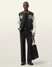 Man wearing black and white varsity jacket with NY logo, black jeans, holding tote bag