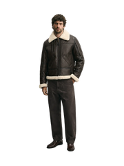 A man wearing a dark brown leather aviator jacket with a cream shearling collar and hem, matching dark brown leather trousers, and brown leather shoes, standing against a light gray studio background.
