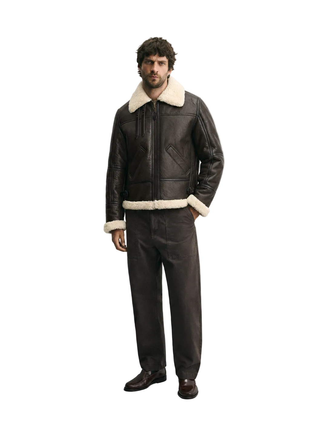 A man wearing a dark brown leather aviator jacket with a cream shearling collar and hem, matching dark brown leather trousers, and brown leather shoes, standing against a light gray studio background.