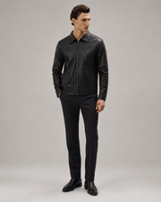 Full-length studio profile of a man wearing the minimalist black leather shirt jacket paired with formal black trousers and black leather dress shoes.