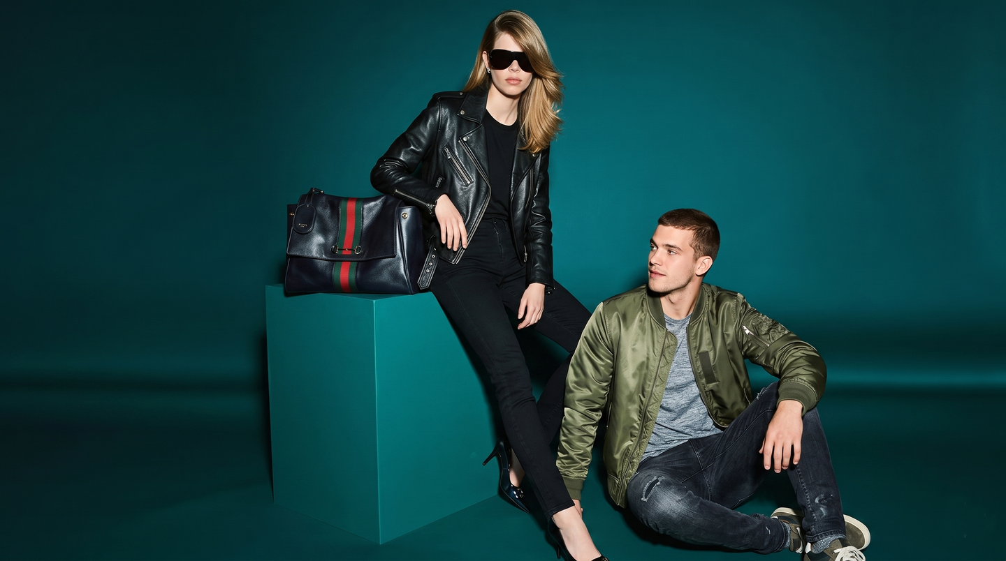 Woman in black leather jacket with designer handbag, man in olive bomber jacket, teal studio background